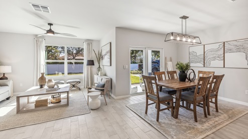 The wooden dining table is set for a family of six. There is a chandelier on top and a paitning on the wall behind. There is an added carpet on top of the tile for comfort. The Dining space is well lit from the patio doors. There are curtains on either side of the patio doors. You can also see the living room from here