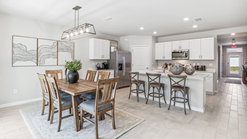 Inside the Cali Model kitchen and Dining room. You can see the entrance hallway from the door. This home includes tiles flooring, stainless steel appliances and white cabinetry with silver fixtures. The Dining space is well lit from the patio doors.