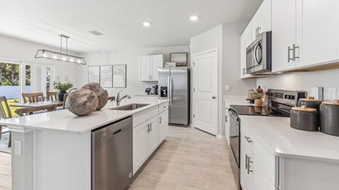 Inside the Cali Model kitchen. This home includes tiles flooring, stainless steel appliances and white cabinetry with silver fixtures. The bar is great for hosting people in the kitchen. You can see the dining room and patio from here. Theres a clear view outside.