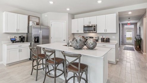 Inside the Cali Model kitchen. You can see the entrance hallway from the door. This home includes tiles flooring, stainless steel appliances and white cabinetry with silver fixtures. The bar is great for hosting people in the kitchen.
