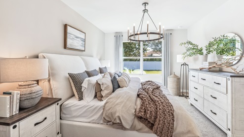 The Master Bedroom is carpeted with grey painted walls. There is a window with a view of the back of the home. There are side tables on either side of the freshly made bed. There is a chandelier above the bed as well.