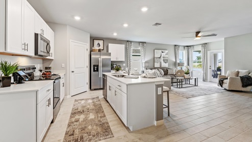 Inside the Lakeside Model Living room and Kitchen. You can see the patio door from here. Floor is tiled and walls are painted grey. All silver/neutral furnishings. The Kitchen is well equipped with all stainless-steal appliances