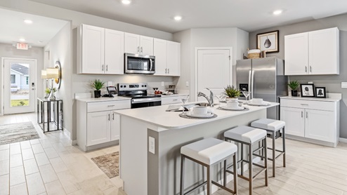 Inside the Lakeside Model Kitchen. You can see the front door from here. Floor is tiled and walls are painted grey. All silver/neutral furnishings. The Kitchen is well equipped with all stainless-steal appliances. There are stools for Bar seating