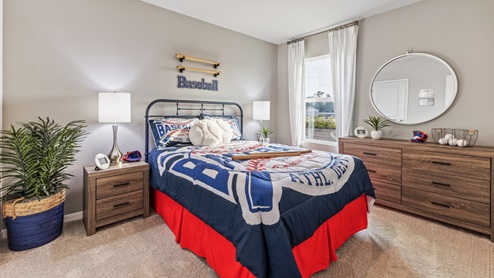 This is a children's bedroom, with a full sized bed, a window with curtains and a baseball theme. There are two side tables, a chest and a plant. Carpeted and painted walls.