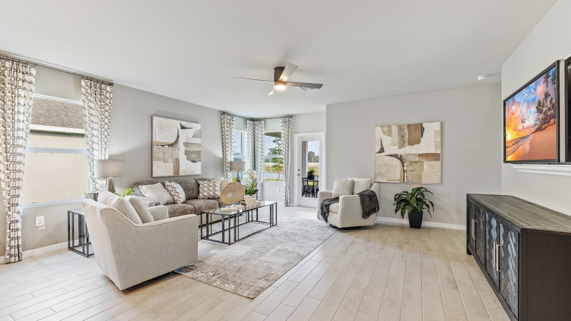 Inside the Lakeside Model Living room. You can see the patio door from here. Floor is tiled and walls are painted grey. All silver/neutral furnishings.  Theres a tv mounted on the wall and a chest underneath