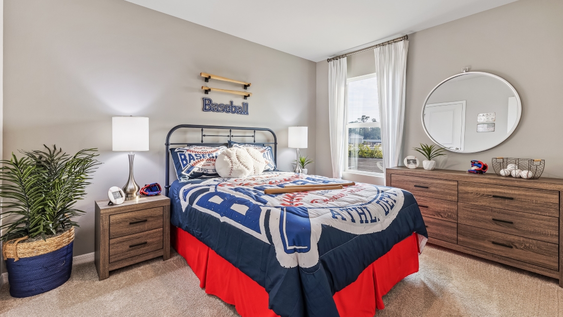 This is a children's bedroom, with a full sized bed, a window with curtains and a baseball theme. There are two side tables, a chest and a plant. Carpeted and painted walls.