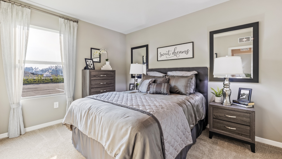 This is a second bedroom, with a full sized bed, a window with curtains and a silver theme. There are two side tables, a chest and a two mirrors on the walls beside the bed. Carpeted and painted walls.