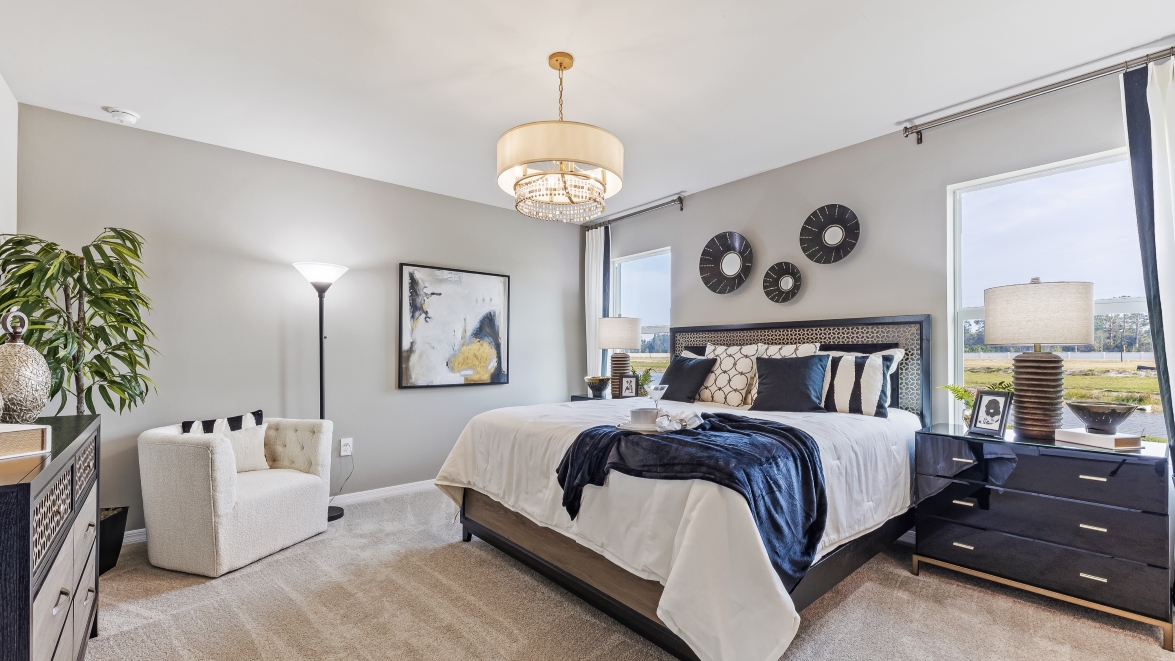 This is a master bedroom, with a king sied bed, 2 windows on either side of the bed with curtains and a navy blue/beige theme. There are two side tables, a chest and a lounge chair across from the bed. Carpeted and painted walls.