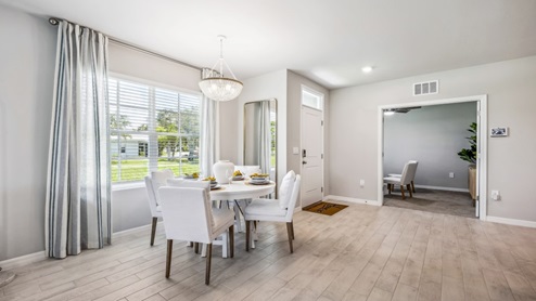 Inside of the Carrington model dining room. Wood tile flooring and white walls. Dining room has a six-person dining table. In the background the living room as well as a window can be seen.