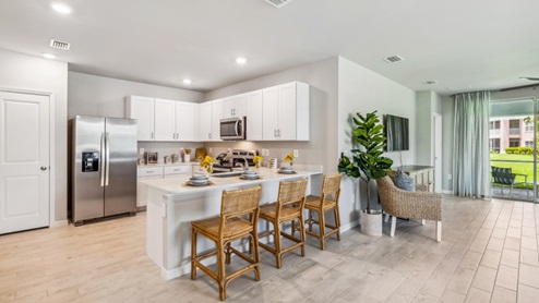 Inside of the Carrington model. Wood tile flooring with white countertops and cabinets with silver hardware. Kitchen island with bar-style seating and stainless-steel appliances. Patio angle
