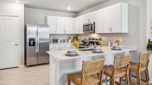 Inside of the Carrington model. Wood tile flooring with white countertops and cabinets with silver hardware. Kitchen island with bar-style seating and stainless-steel appliances. Bar angle