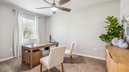 Inside of the Carrington model. Carpet flooring with off-white walls. Room contains a desk, two chairs, and dresser with various vases and a plant. Room contains window.