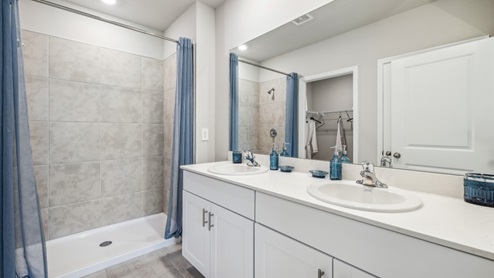 Inside of the Carrington model bathroom one. Wood tile flooring with off-white walls. Double vanity with white countertops and cabinets with silver hardware. Shower has grey tile and silver hardware.