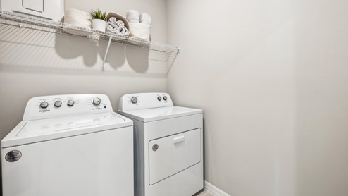 Inside of the Carrington model laundry room. Room contains a white washer and dyer as well as a white shelf with a hanger on it over the washer and dryer.