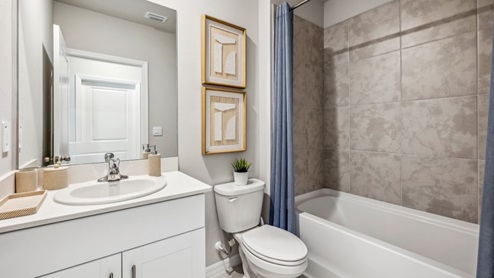 Inside of the Carrington model bathroom one. Wood tile flooring with off-white walls. Singular vanity with white countertops and cabinets with silver hardware. Shower with tub has grey tile and silver hardware.