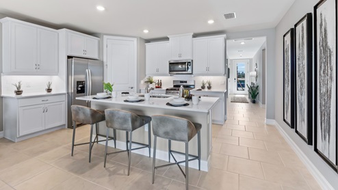 Inside of the Cali model kitchen. Tile flooring and white walls. Kitchen has white countertops and cabinets with silver hardware, kitchen island with bar-style seating, and stainless-steel appliances.