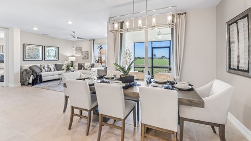 Inside of the Cali model dining room. Tile flooring and white walls. Dining room has a six-person dining table with hanging light fixture above it. Kitchen can be seen in the background.