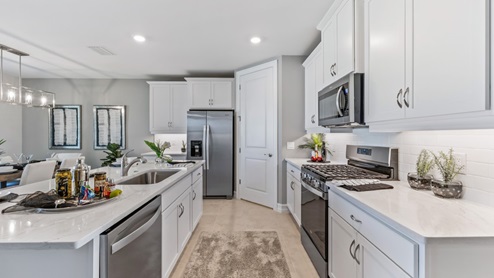 Inside of the Cali model kitchen. Tile flooring and white walls. Kitchen has white countertops and cabinets with silver hardware, kitchen island with bar-style seating, and stainless-steel appliances.
