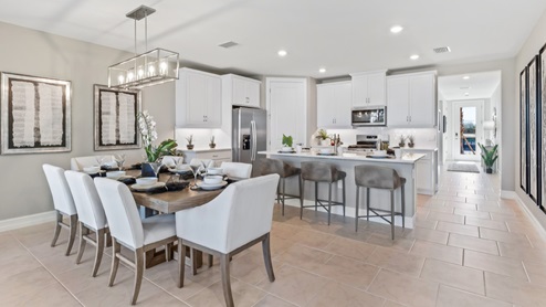 Inside of the Cali model dining room. Tile flooring and white walls. Dining room has a six-person dining table with hanging light fixture above it. Kitchen can be seen in the background.