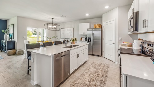 Inside of the Cali model kitchen. Wood tile floors with teal walls. Kitchen contains white countertops and cabinets with silver hardware. There is a kitchen island with bar-style seating. Kitchen appliances are stainless-steel.