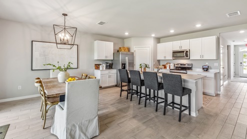 Inside of the Cali model dining area. Wood tile flooring and teal walls. Dining area contains a six-person dining table with a hanging light fixture above it. In the background a sliding glass door that leads to the back patio can be seen as well as an open area that contains the living room furniture.