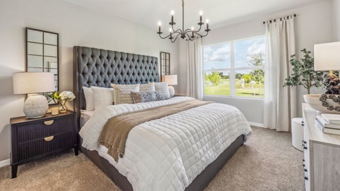 Inside of the Cali model bedroom. Crème carpet with off-white walls. Room contains a bed with nightstands on either side as well as a plant in the corner of the room.