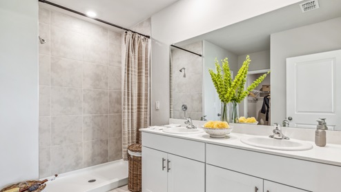 Inside of the Cali model bathroom. Shower has grey tile and silver hardware. Room contains a dual vanity with white countertops and cabinets with silver hardware. Bathroom also contains a closet.
