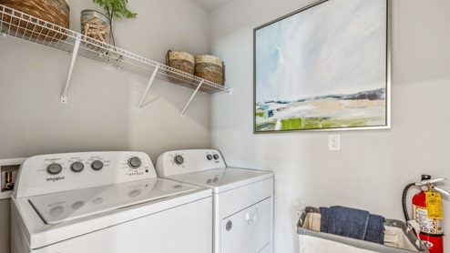 Inside of the Cali model laundry room. Room contains a white washer and dryer side by side. There is also a shelf above the washer and dryer with hangers on it.