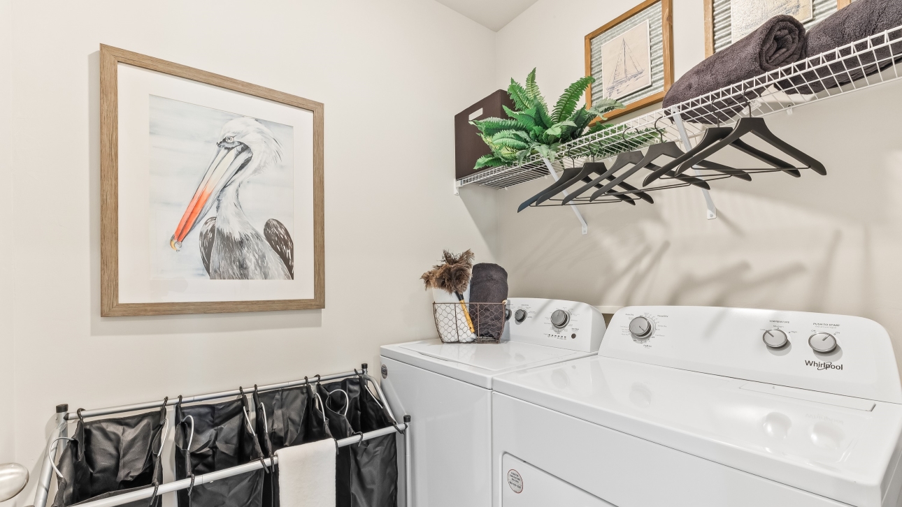 Inside of the Cali model laundry room. Room contains a white washer and dryer side by side. There is also a shelf above the washer and dryer with hangers on it.