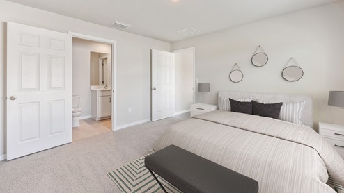 Inside of the Freeport bedroom. White carpet with creme walls. Bedroom has a connected bathroom. Room contains a large bed with grey sheets as well as two small nightstands on either side of bed.