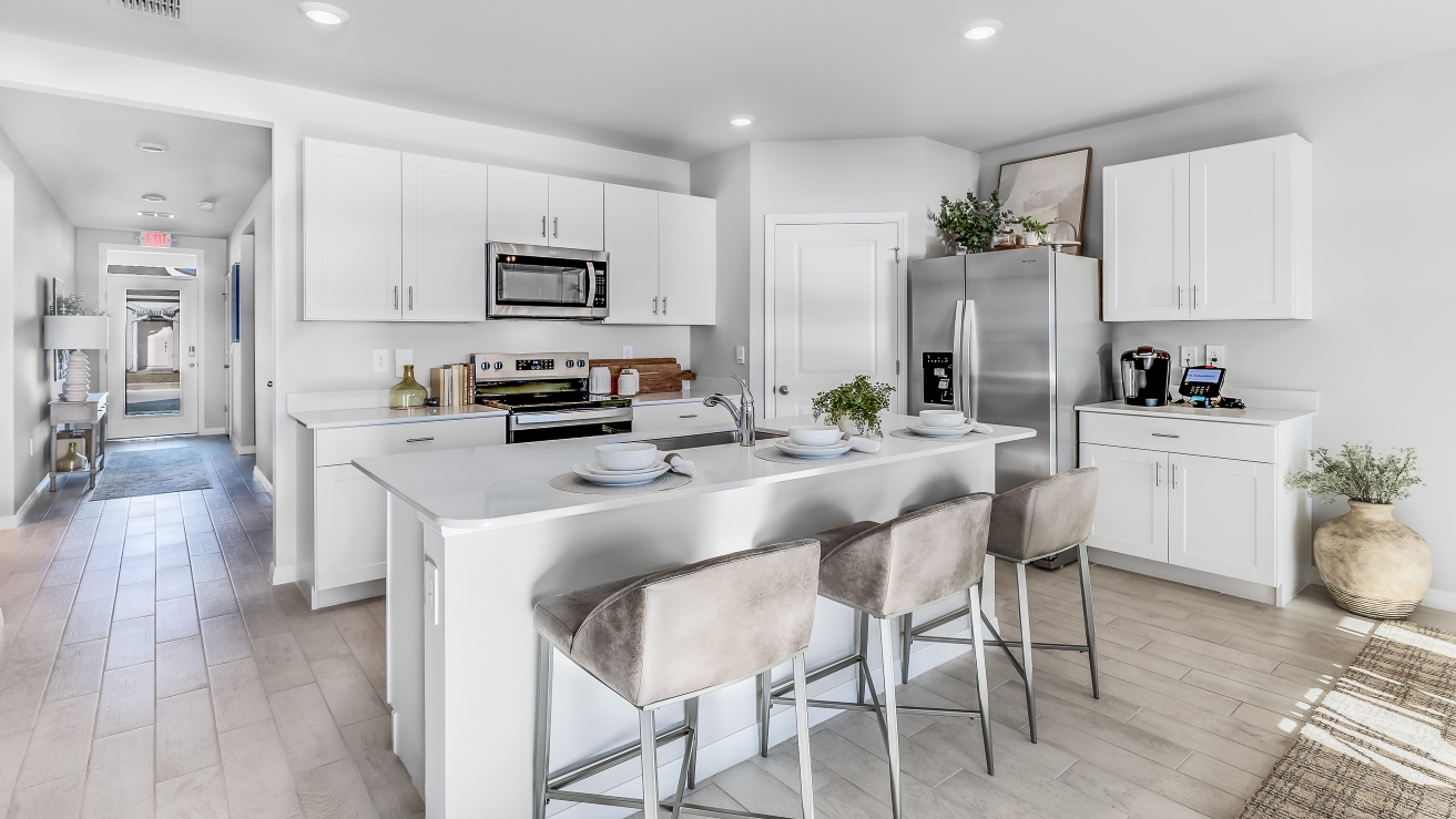 Inside the Cali Model kitchen. You can see the entrance hallway from the door. This home includes tiles flooring, stainless steel appliances and white cabinetry with silver fixtures. The bar is great for hosting people in the kitchen.