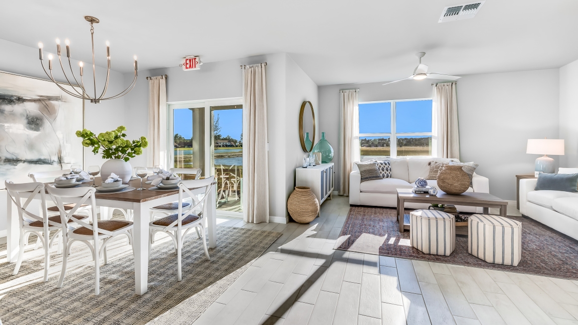 The wooden dining table is set for a family of six. There is a chandelier on top and a paitning on the wall behind. There is an added carpet on top of the tile for comfort. The Dining space is well lit from the patio doors. There are curtains on either side of the patio doors. You can also see the living room from here
