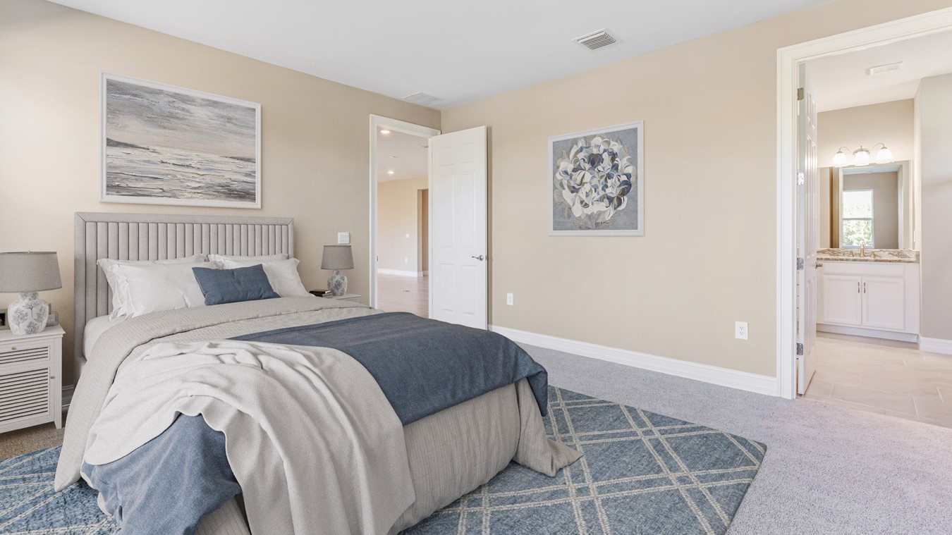 Inside the Kellen model Master Bedroom with view to the bathroom and door to living room. Walls are tan and carpet is gray. Bedding is gray/blue. There are two side tables on either side of the bed with lamps on each.