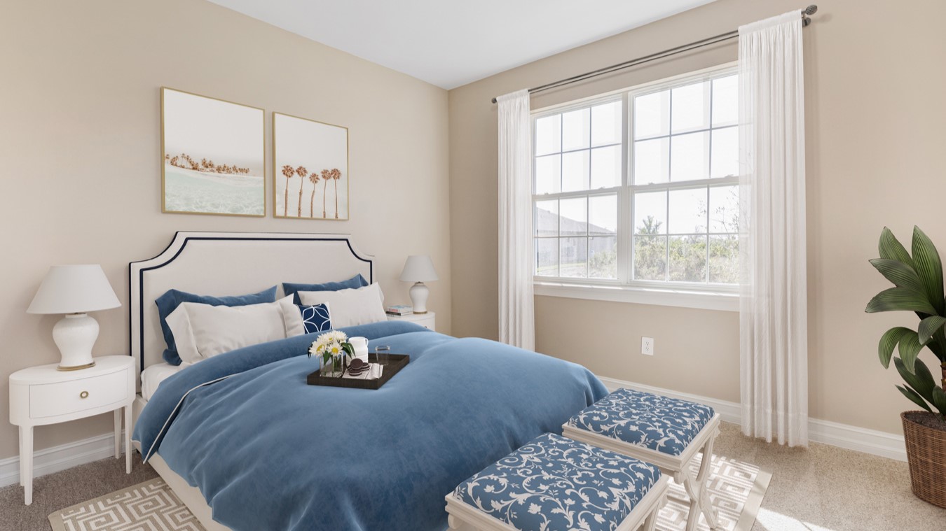 Inside the Kellen model second Bedroom with view of the large double paned window with 3x4 grid. Walls are tan and carpet is beige.  Bedding is creme/blue. There are two white side tables on either side of the bed with lamps on each and two seats at the foot of the bed.