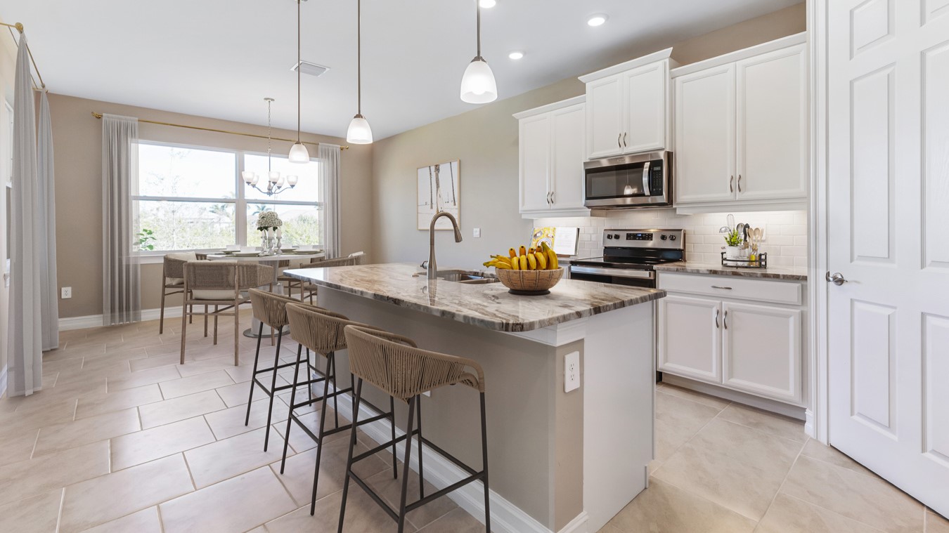 Inside the Kellen model kitchen area, appliances are brand new black and silver. There is a kitchen Island and Bar. Pantry and cabinetry is white with silver fixings. You can see the dining area with a view outside a large 2x2 window with curtains on either side.