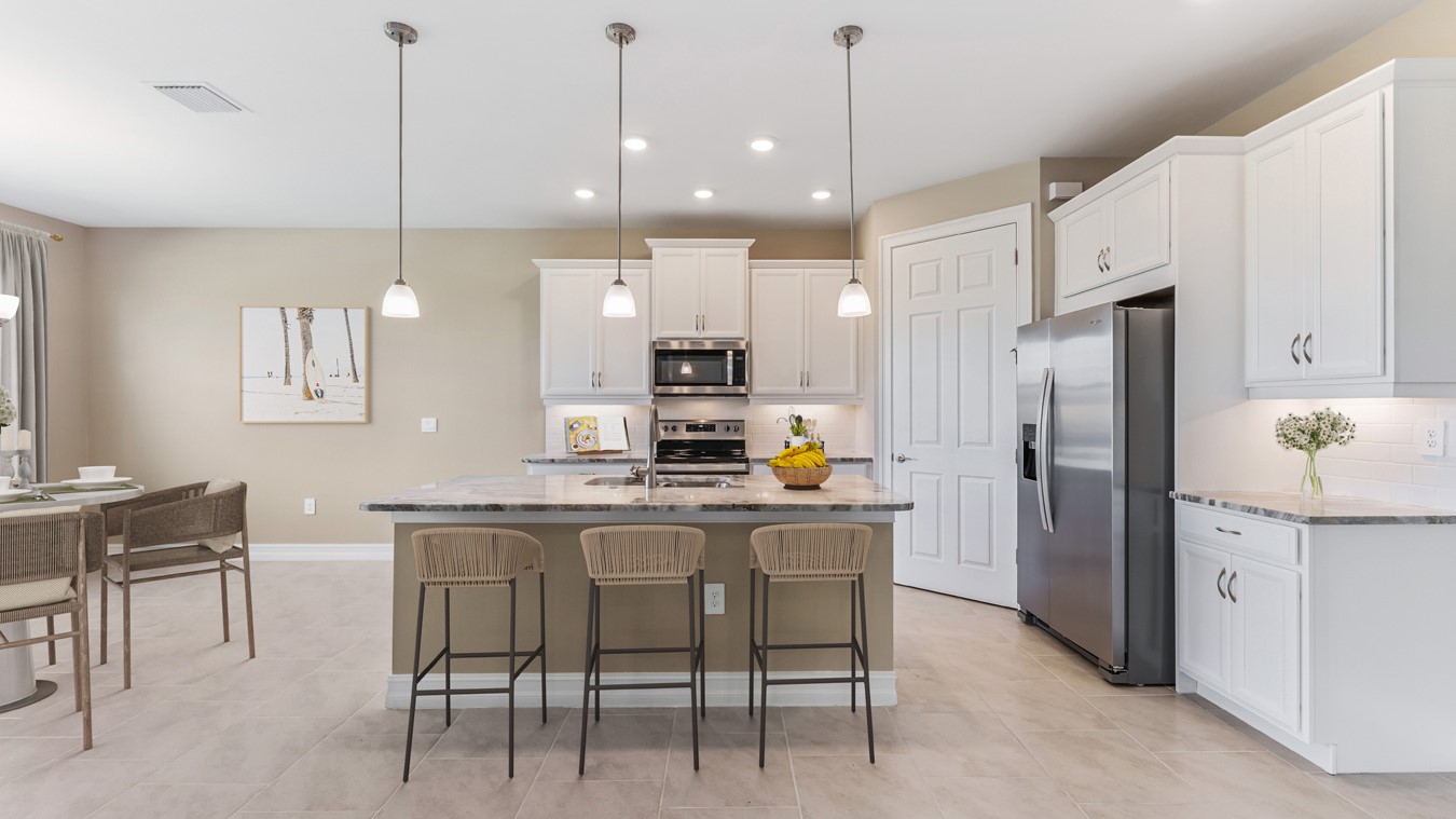 Inside the Kellen model kitchen area, appliances are brand new black and silver. There is a kitchen Island and Bar. Pantry and cabinetry is white with silver fixings. You can see partial of the dining area. This is more cabinetry and the refrigerator.