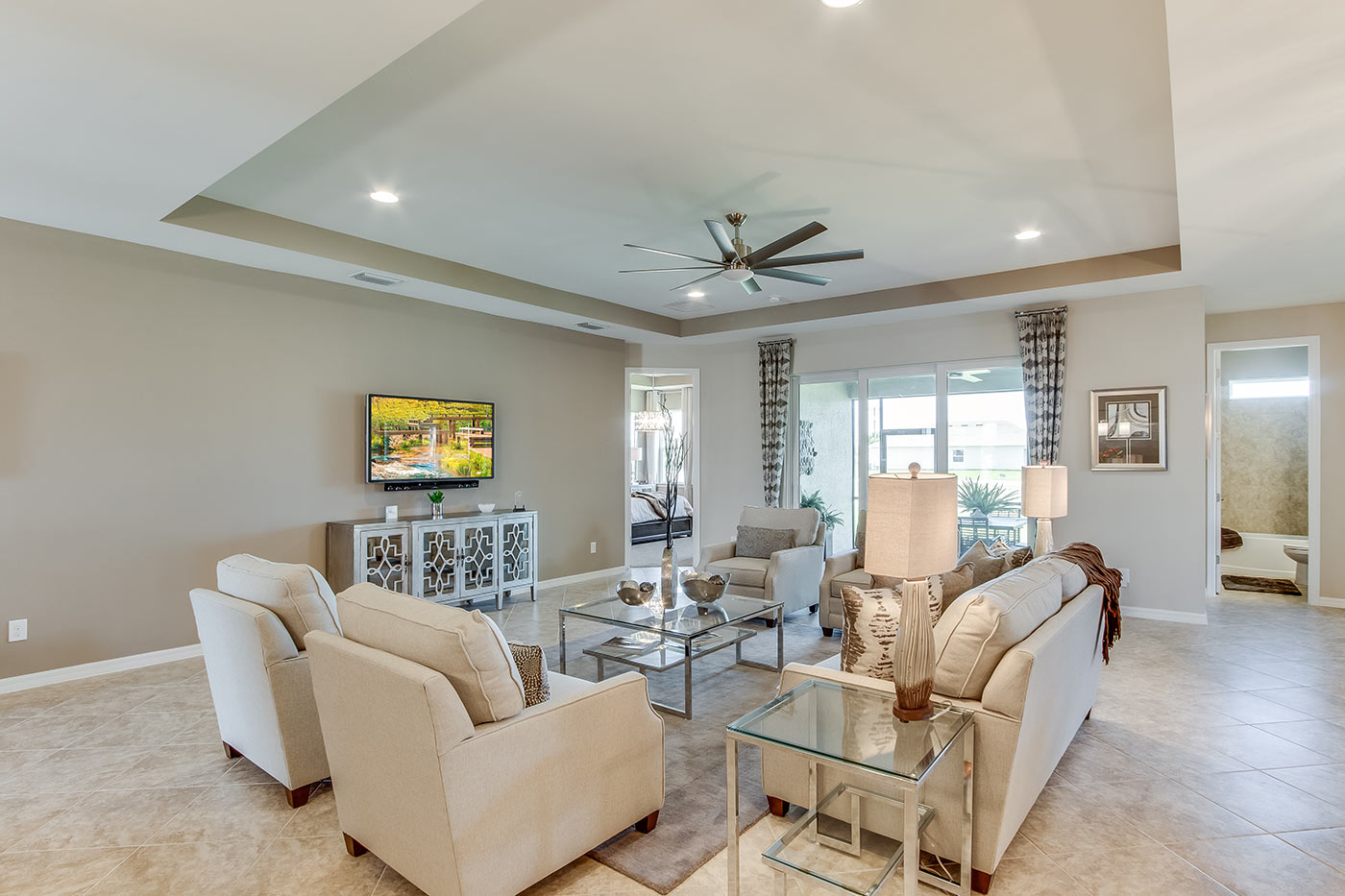 Inside of the Destin model living room. Tile flooring and off-white walls. Living room contains a couch, four armchairs, coffee table, side table, TV, and TV stand. In the background the dining area can be seen.