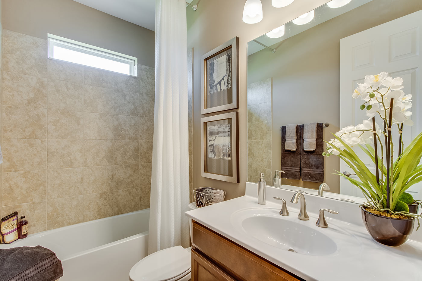 Inside of the Destin model bathroom. Off-white walls. Single vanity with white counters and wooden cabinets with silver hardware. Shower-tub combo has crème tile and white tub with silver hardware.