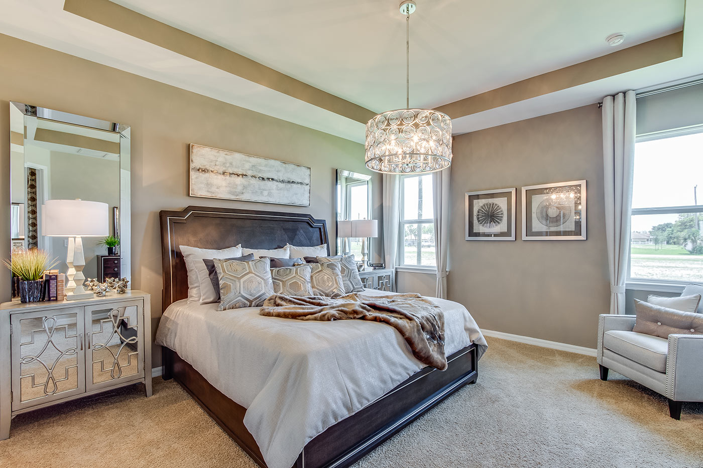 Inside of the Destin model bedroom two. Crème carpet and off-white walls. Bedroom contains a bed with nightstands on either side of the bed and armchair. Room also has two windows.