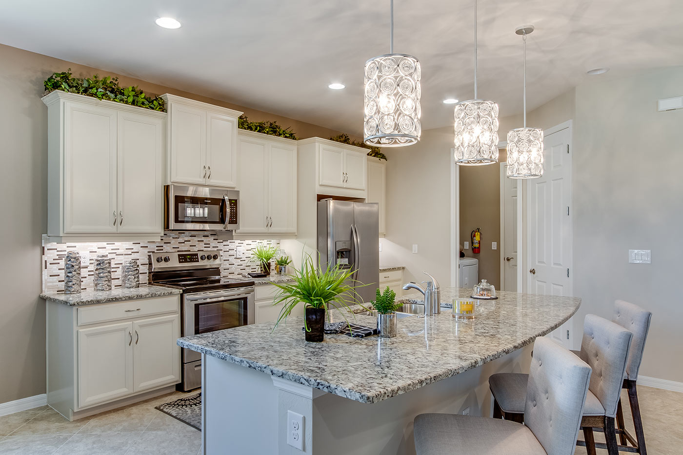 Inside of the Destin model kitchen. Tile flooring and off-white walls. Kitchen has grey countertops with white cabinets and silver hardware. Bar-style kitchen island.