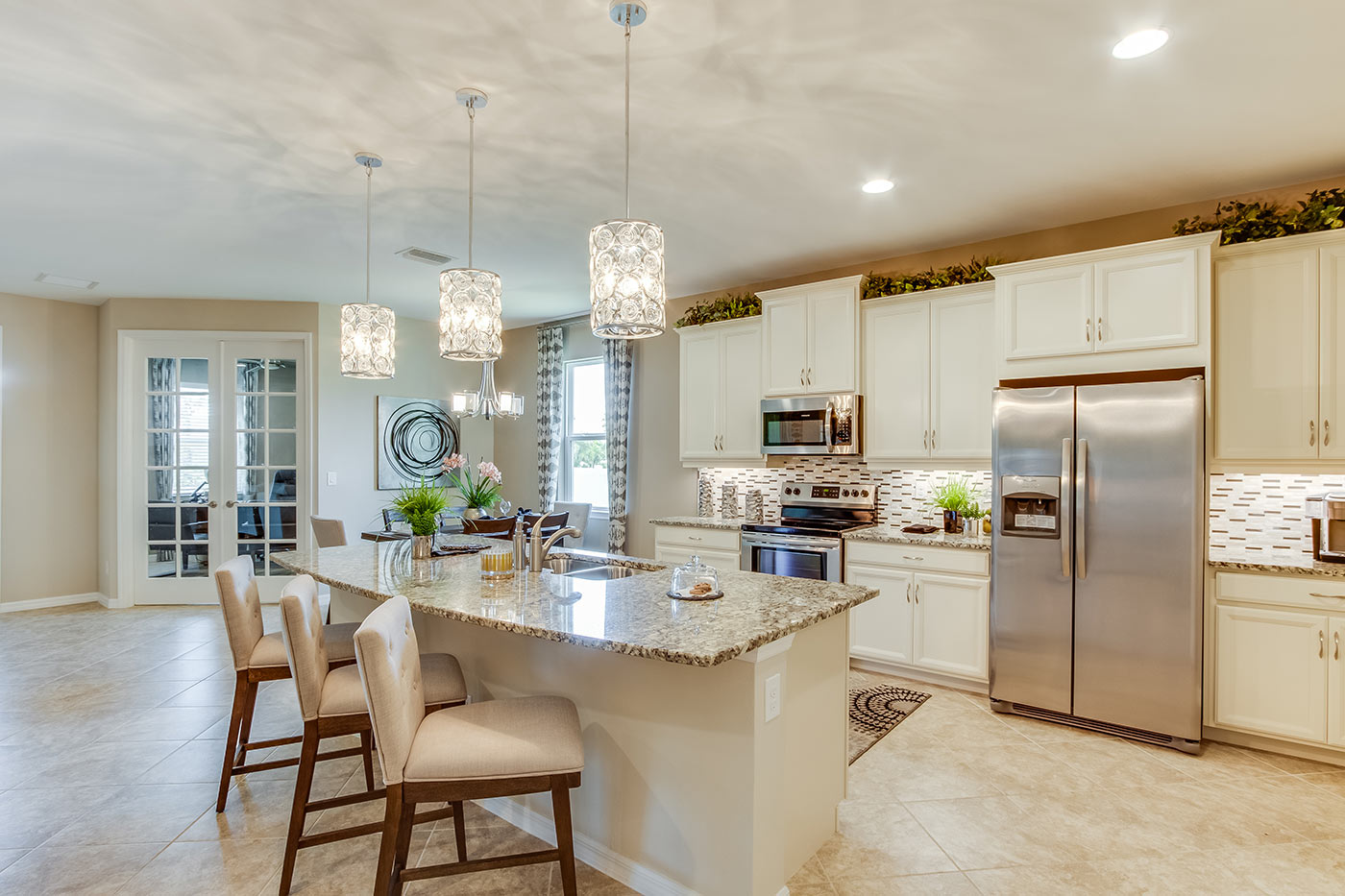 Inside of the Destin model kitchen. Tile flooring and off-white walls. Kitchen has grey countertops with white cabinets and silver hardware. Bar-style kitchen island. In the background the den can be seen.