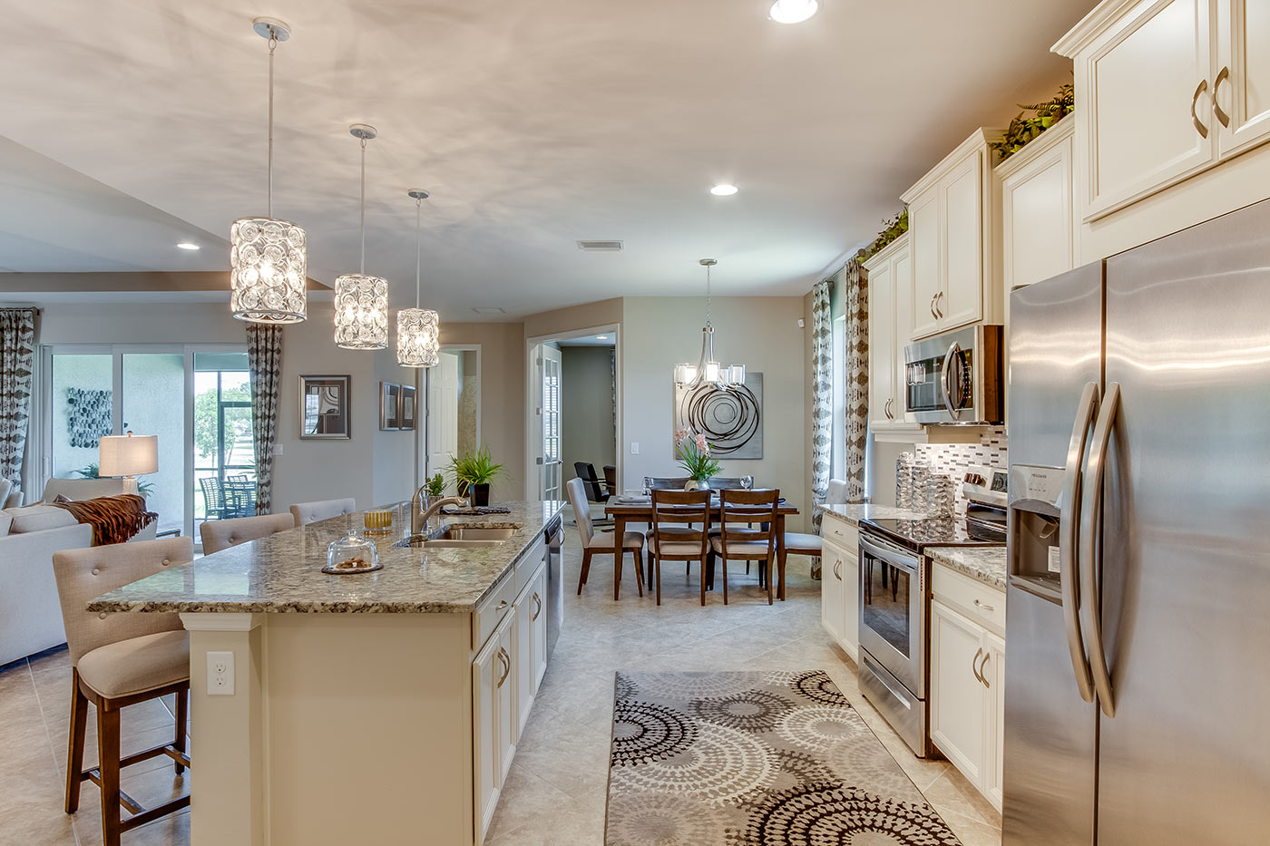 Inside of the Destin model kitchen. Tile flooring and off-white walls. Kitchen has grey countertops and white cabinets and silver hardware. Bar-style seating. In the background the dining area can be seen.