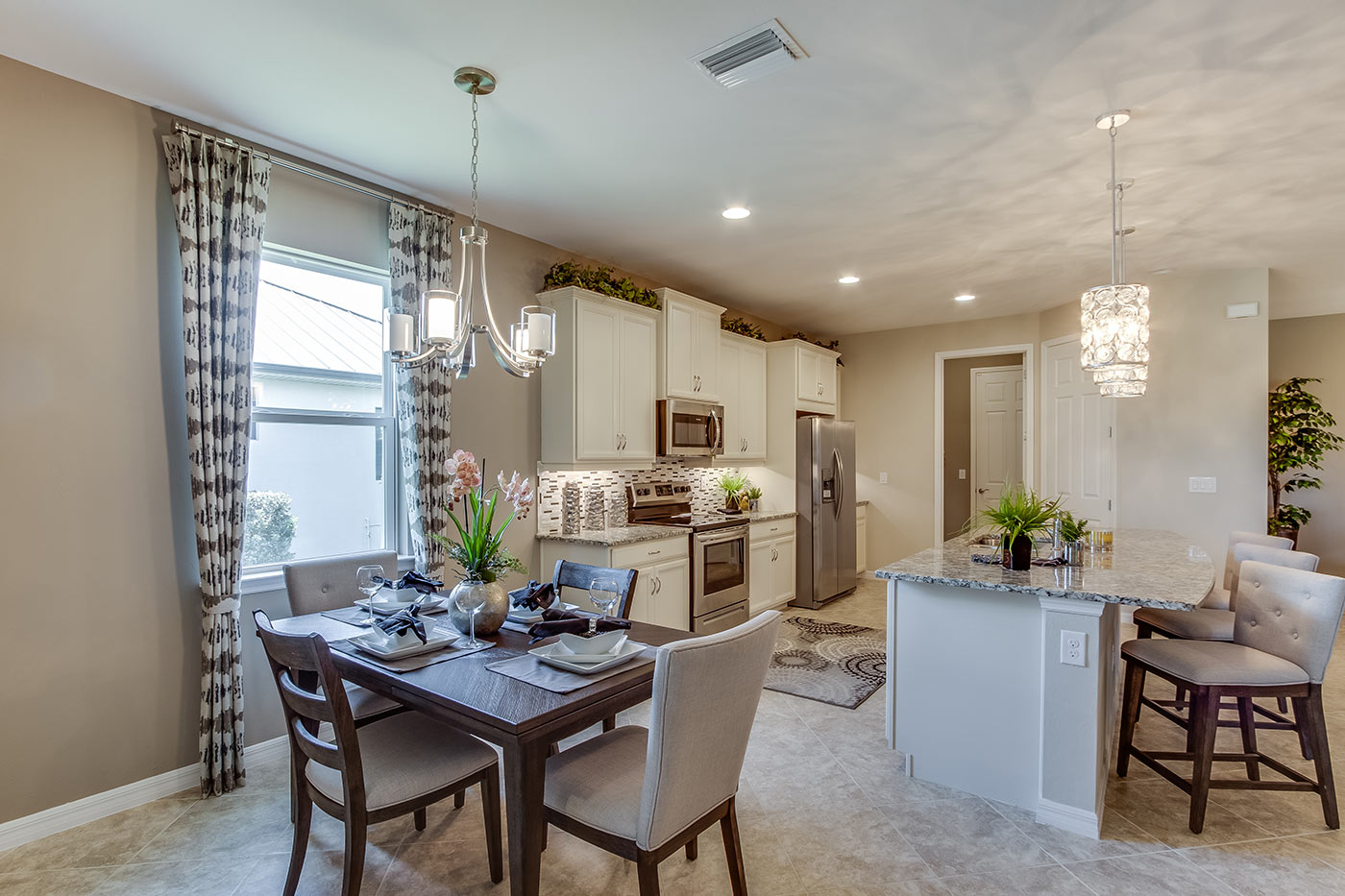 Inside of the Destin model dining room. Tile flooring and off-white walls. Dining area has a four person dining table with a hanging light fixture. Kitchen can be seen in the background.