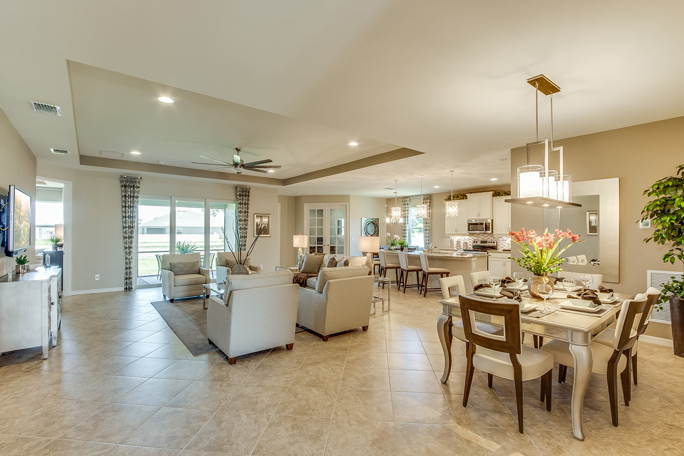 Inside of the Destin model living room. Tile flooring and off-white walls. Living room contains a couch, four armchairs, coffee table, side table, TV, and TV stand. In the background the kitchen, dining area, den, and sliding glass door that leads to back patio can be seen.