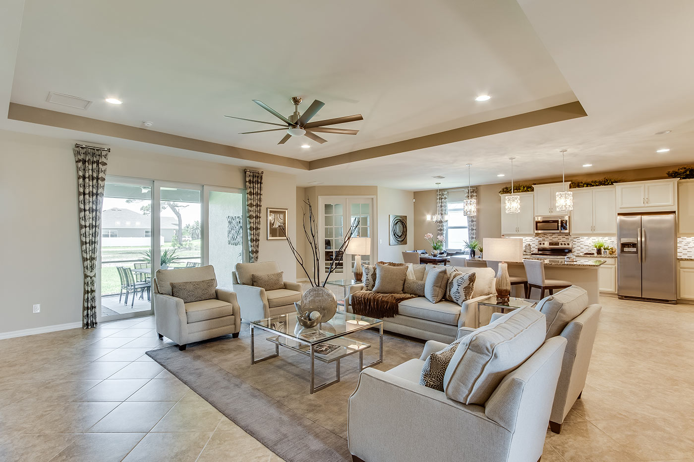 Inside of the Destin model living room. Tile flooring and off-white walls. Living room contains a couch, four armchairs, coffee table, side table, TV, and TV stand. In the background the kitchen, den, and sliding glass door that leads to back patio can be seen.