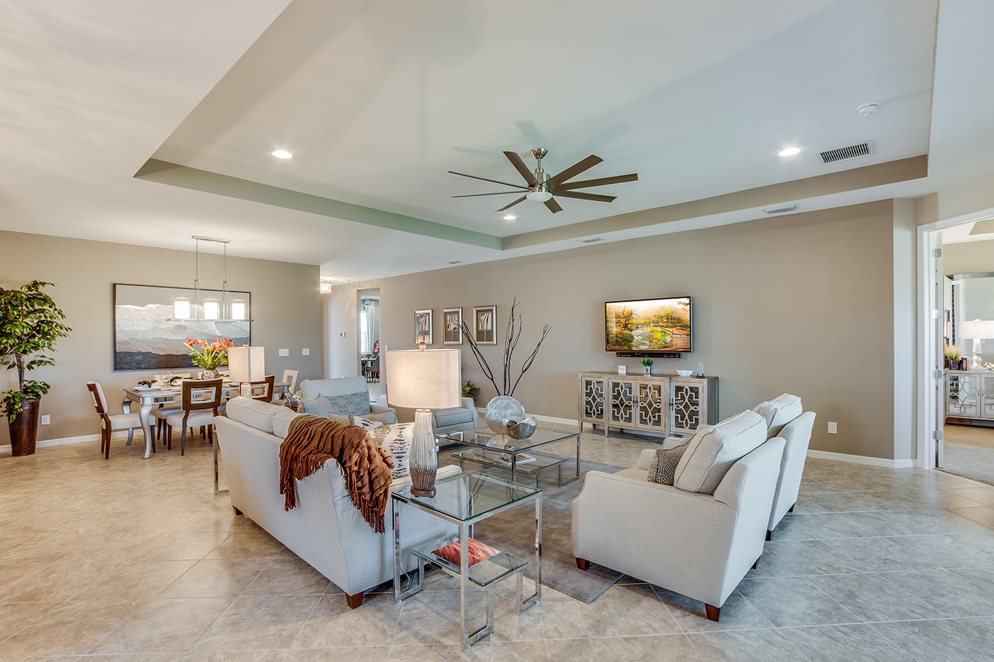 Inside of the Destin model living room. Tile flooring and off-white walls. Living room contains a couch, four armchairs, coffee table, side table, TV, and TV stand. In the background the dining area can be seen.