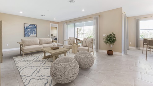 Inside the Kellen model Great Room from infront of the ottomans and seating arrangment. There is also a the large couch. You can see the patio sliding doors, and a slight area of the dining from here. Tiled throughout. You can see to the master bedroom.