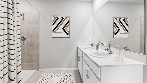 Inside of the Aria model bathroom. Wood tile floor with grey walls. White countertops with stainless steel hardware. Bathroom contains a double vanity with white cabinets as well as a shower with dark grey tile.