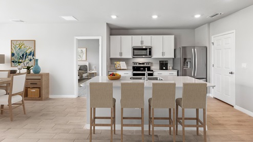 Inside of the Aria kitchen. Wooden tile floor and off-white walls. Island with white countertops and bar-style seating. White cabinets with silver hardware. Stainless steel refrigerator, microwave, stove, and sink.