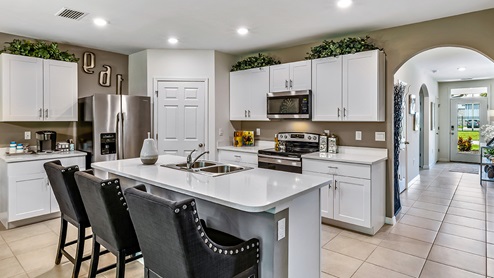 Inside of the Cali model kitchen. Tile flooring and white walls. Kitchen has white countertops and cabinets with silver hardware, kitchen island with bar-style seating, and stainless-steel appliances.